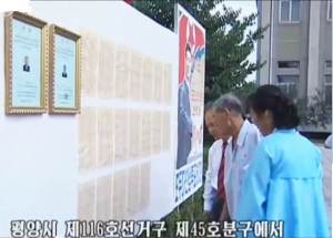 Voters look at the stand featuring the DPRK election trifecta (from right to left): the WPK’s election propaganda poster, the LEC-approved lists of registered voters (19 pages by 50 names = 950 voters), and the certified notices of the deputy candidates registered at the electoral district No. 116, sub-constituency No. 45, at the Pyongyang Textile Machinery Plant, in Songyo district, Pyongyang, on July 19, 2015. Photo: KCTV.