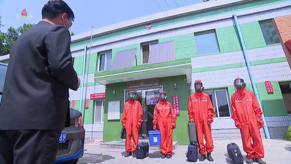 Four workers are standing in a line wearing red protective equipment and gas masks